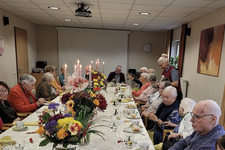 Ältere Menschen sitzen an einem langen, festlich gedeckten Tisch mit Blumenarrangements und Kerzen in einem Raum mit Deckenbeleuchtung und einer Uhr an der Wand