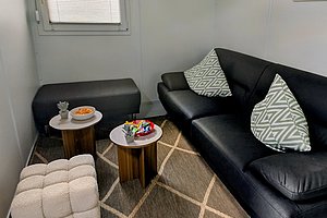 Kleines Wohnzimmer mit schwarzem Ledersofa, zwei gemusterten Kissen, zwei kleinen runden Beistelltischen, einem gepolsterten Hocker und einem Teppich mit geschwungenem Linienmuster