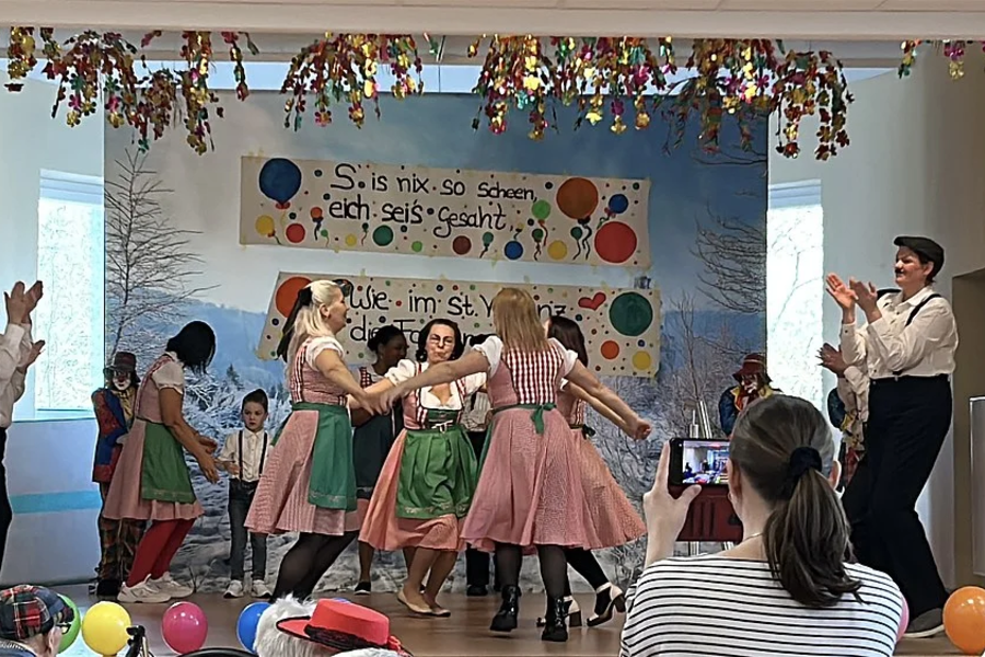 Gruppe von Menschen in Trachten tanzt auf einer Bühne, Zuschauer filmen mit Smartphone, bunte Luftballons und Girlanden als Dekoration