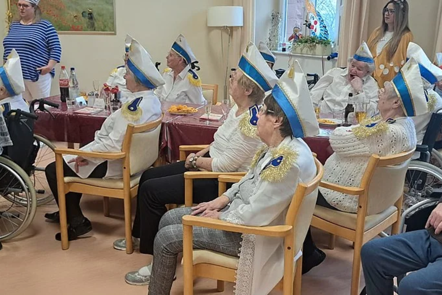 Mehrere ältere Menschen in weißen Uniformen mit blauen Mützen sitzen in einem Raum auf Stühlen, im Hintergrund stehen zwei Frauen, eine davon in einem Rollstuhl