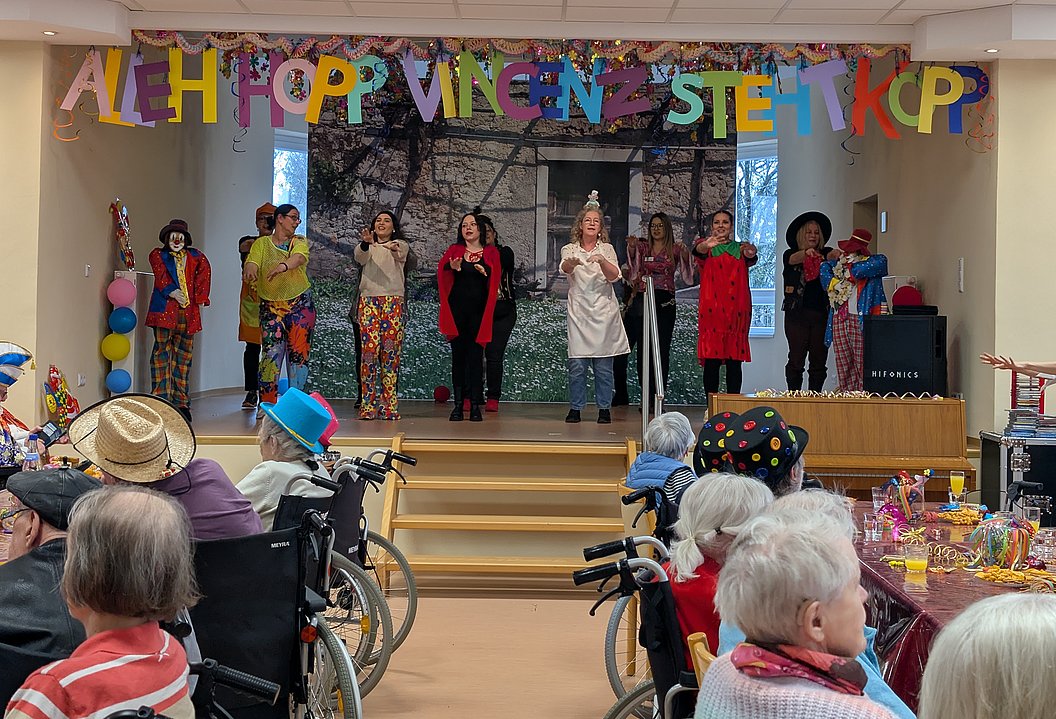 Bunt geschmückter Saal mit Aufführung, Senioren im Vordergrund, fröhliche Stimmung und bunte Dekoration.