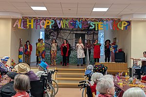 Bunt geschmückter Saal mit Aufführung, Senioren im Vordergrund, fröhliche Stimmung und bunte Dekoration.