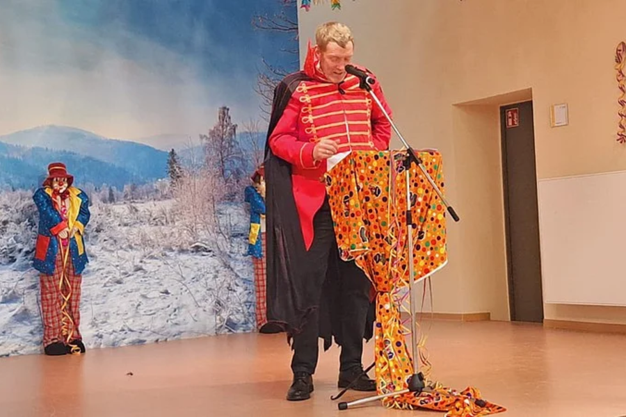 Person in rotem Kostüm mit Umhang steht an mit buntem Stoff bedecktem Rednerpult in einem Raum mit winterlicher Landschaft als Wandbild und zwei weiteren kostümierten Personen im Hintergrund