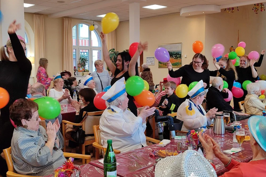 Mehrere ältere Menschen und Betreuer in einem festlich geschmückten Raum halten bunte Luftballons und klatschen an Tischen mit Getränken und Dekoration