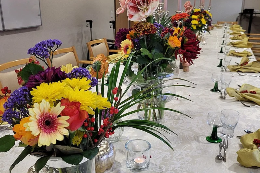 Langer festlich gedeckter Tisch mit mehreren bunten Blumenarrangements und brennenden Kerzen in einem Raum mit Stühlen und Whiteboard