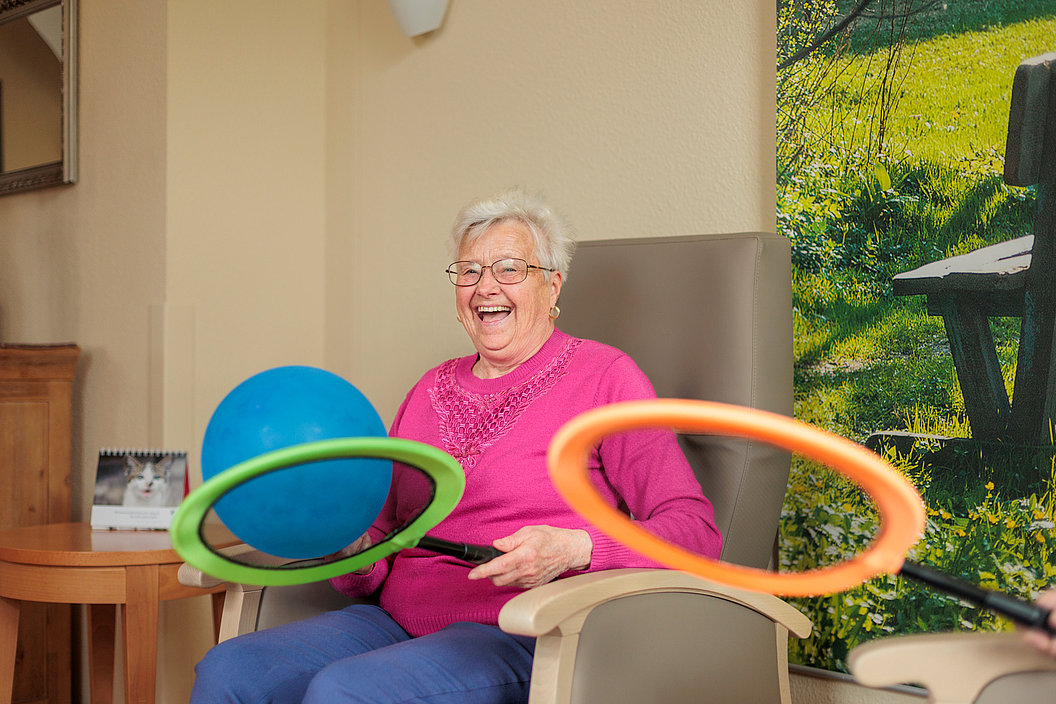 Ältere Frau mit Brille sitzt lachend im Sessel, hält bunte Ringe und einen blauen Ball.