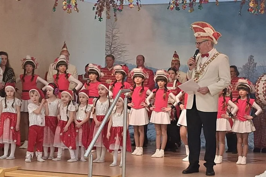 Gruppe von Kindern in roten und weißen Kostümen auf einer Bühne mit einem Mann in weißem Anzug und rotem Hut davor
