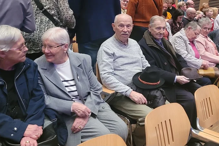 Mehrere ältere Menschen sitzen in einer Reihe auf Holzbänken in einem Innenraum, einige stehen im Hintergrund.