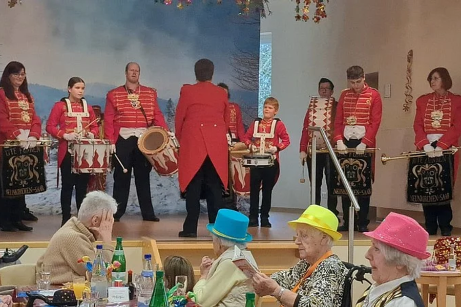 Gruppe von Musikern in roten Uniformjacken mit Trommeln und Blasinstrumenten auf einer Bühne vor sitzendem Publikum mit bunten Hüten