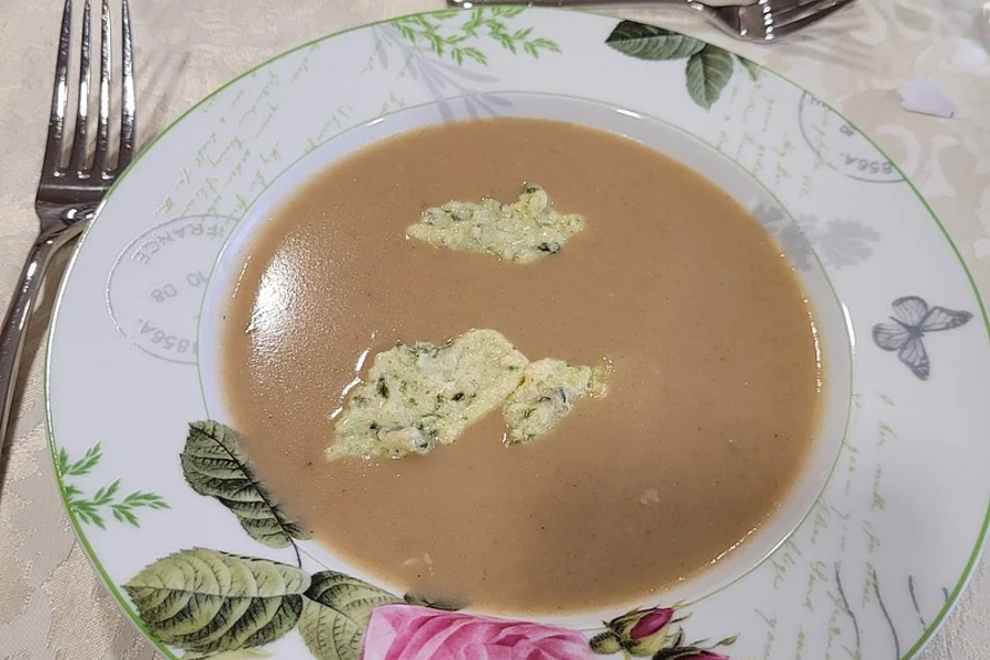 Teller mit brauner Suppe und zwei Klecksen grüner Creme auf einem weißen Tisch mit Besteck und Gläsern