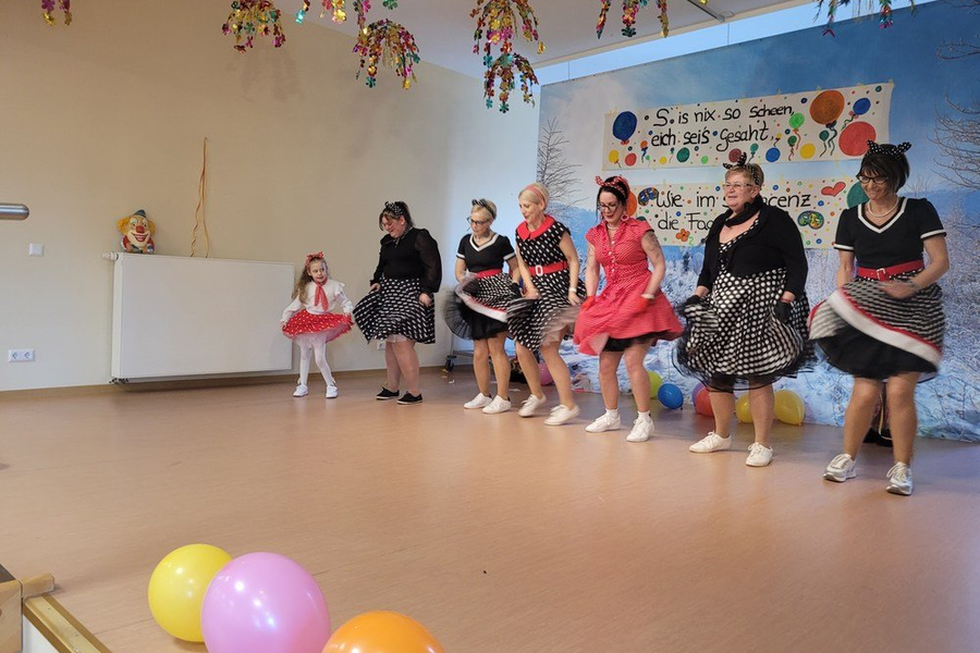 Sieben Frauen und ein kleines Mädchen tanzen in einem Raum mit bunten Luftballons und einer Wanddekoration mit Text und Blumen.