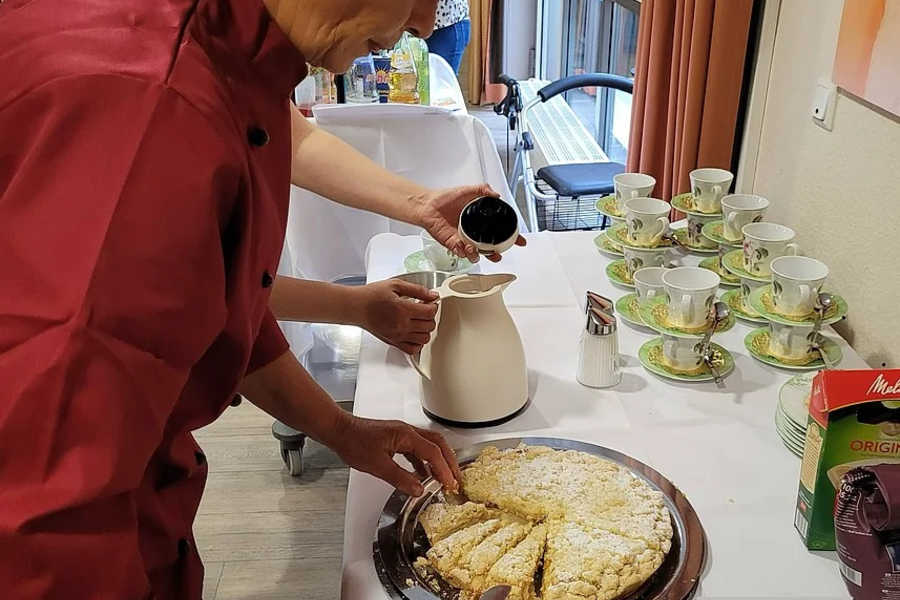 Person gießt aus einem weißen Kännchen Flüssigkeit in eine Tasse neben einem Tisch mit mehreren Tassen und einem großen Kuchen auf einem Tablett