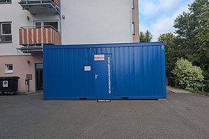 Blauer Container mit Tür und Aufschrift vor einem Wohnhaus mit Balkonen und Bäumen im Hintergrund