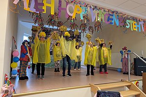 Bunte Aufführung mit Menschen in Regenponchos, Menschen die tanzen und jubeln, vor festlicher Dekoration.