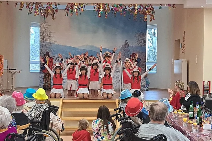 Kinder in roten Kostümen stehen auf einer Bühne und führen eine Tanzvorführung vor einem Publikum aus älteren Menschen in einem Saal auf
