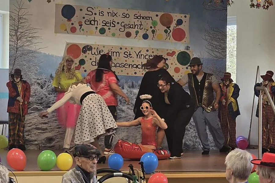 Bühnenauftritt mit mehreren Personen in bunten Kostümen vor einer Wand mit handgemalten Schildern und Luftballons auf der Bühne und im Publikum.