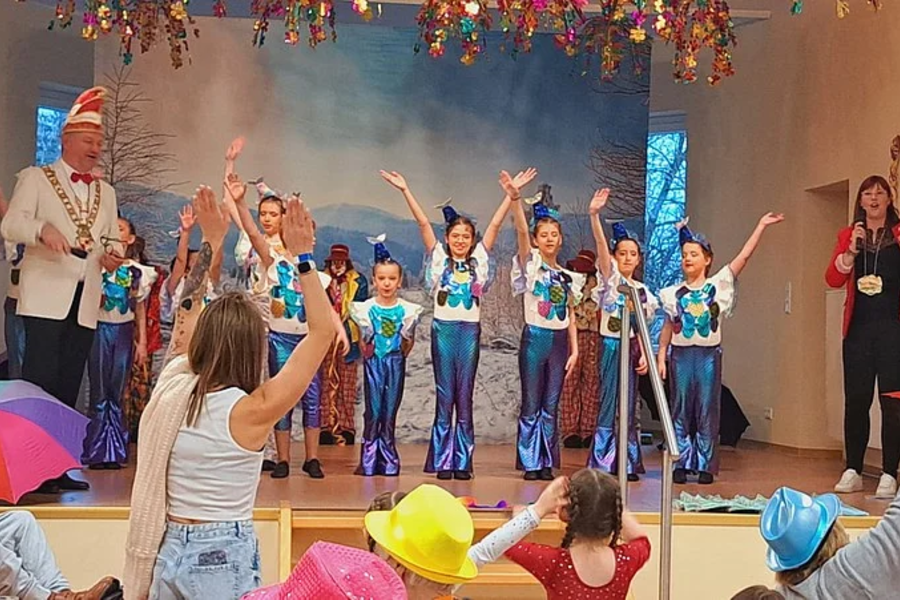 Kinder in bunten Kostümen stehen auf einer Bühne und winken, davor sitzen Zuschauer mit Hüten und Regenschirmen in einem Saal